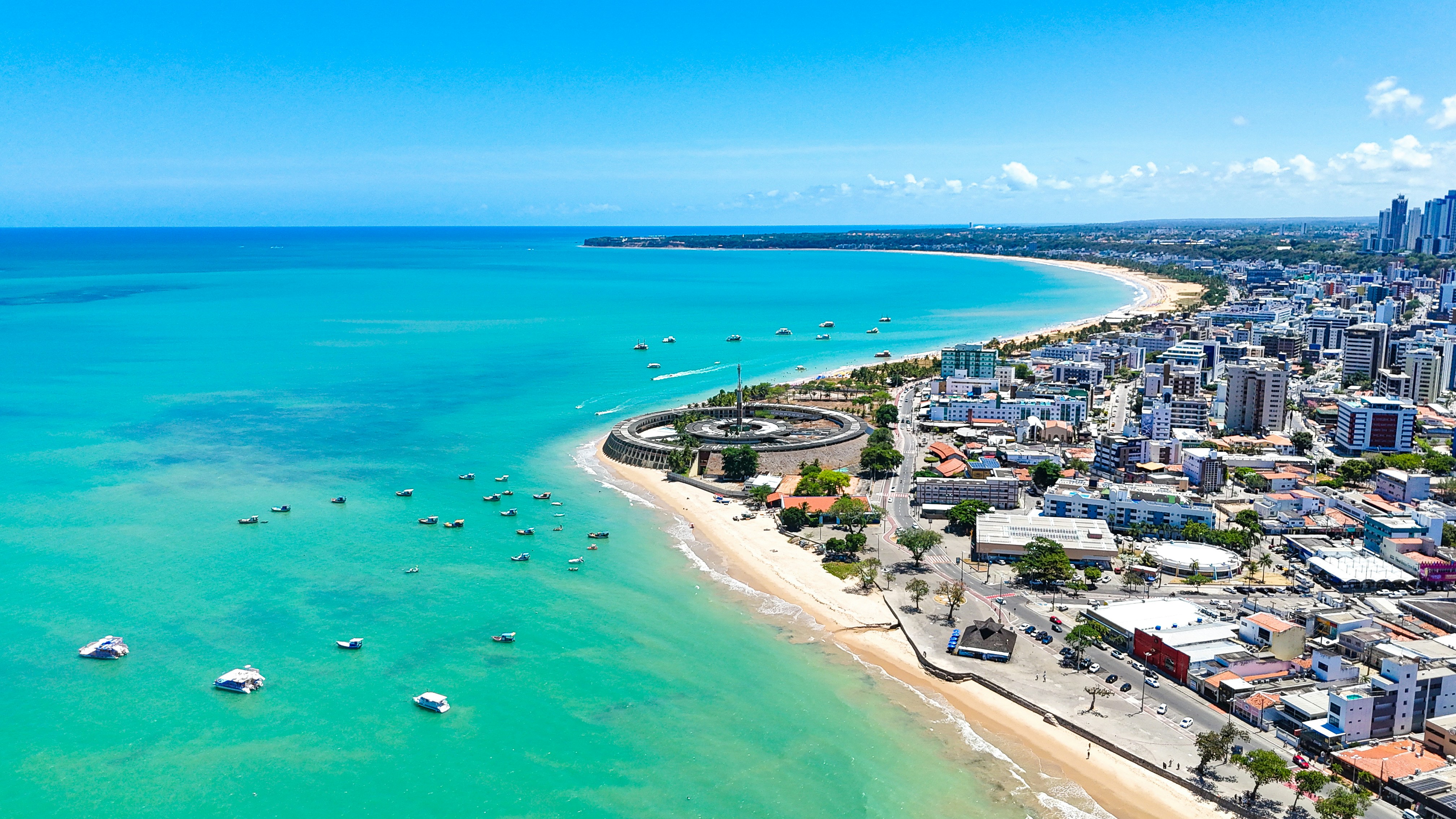 Vista aérea da orla de João Pessoa, com a praia, os barcos e a cidade acompanhando a costa